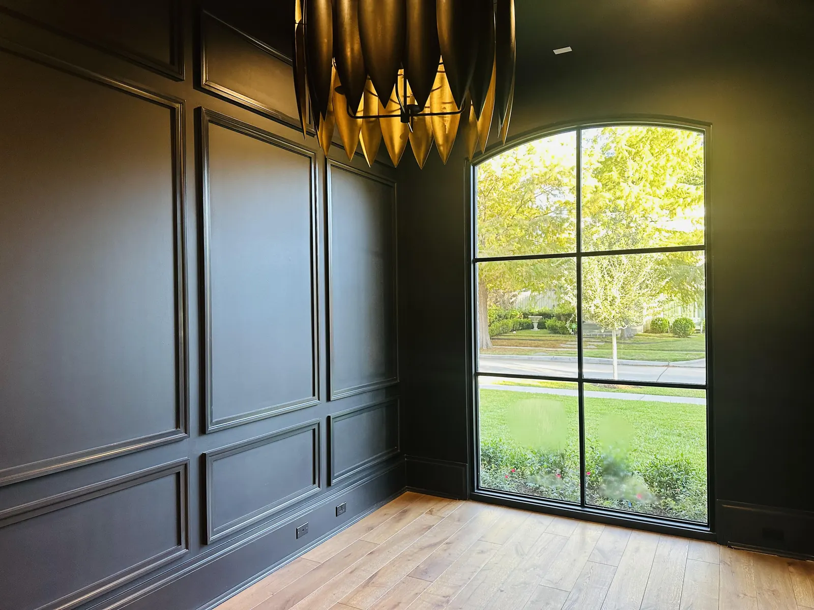 Dining room with dark painted wall panels, a large arched window and a gold chandelier